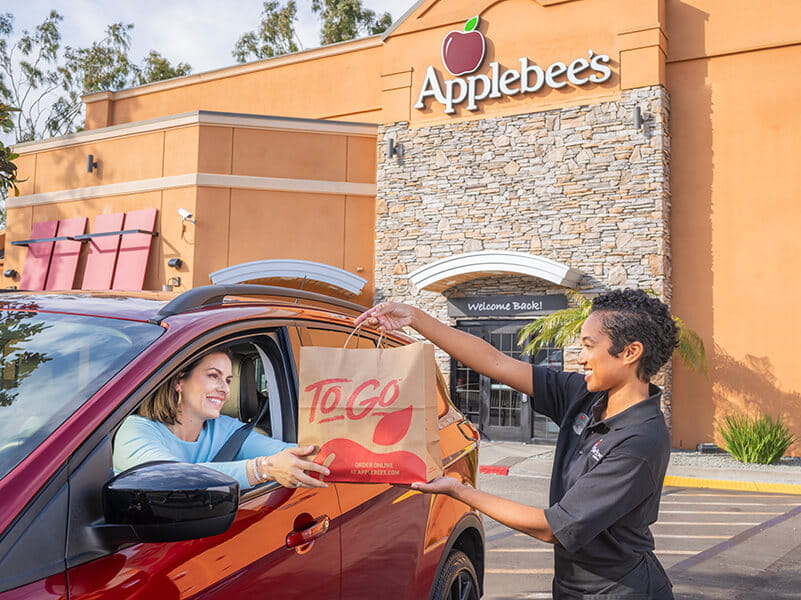 Image from the 2000s era shwoing an Applebee's female team member handing a carside-to-go order to a female in her vehicle